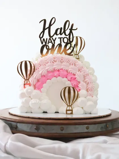 Celebrating a special milestone with a 'Half Way to One' cake. This adorable half-cake design features a pink and white rainbow made of piped frosting clouds and cute hot air balloon toppers.