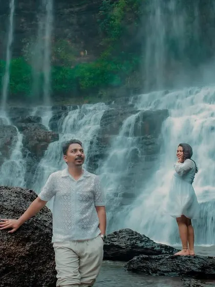 An artistic, moody shot of the couple at the waterfall. They stand apart, looking in different directions, creating a cinematic and thought-provoking image.