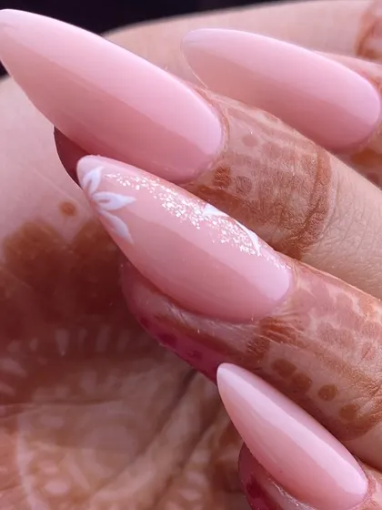 A close-up of a delicate design on almond nails, featuring a soft pink base with a subtle white floral accent and a sprinkle of glitter. This is perfect for an engagement or minimalist bridal look.