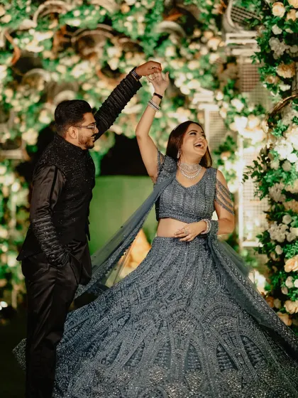 A happy couple enjoying their sangeet night. The bride's twirling grey lehenga and the groom's classic black sherwani are perfectly complemented by the lush green floral backdrop I designed for their photo corner.