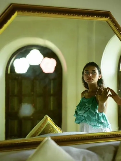 A creative indoor portrait showing a woman's reflection in a large, ornate mirror. Another person's hand enters the frame to touch the mirror, adding a layer of mystery and narrative to the scene.