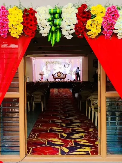 A traditional and welcoming entrance to a wedding hall. The doorway is framed with bright red drapes and a colorful flower toran, a classic element in Indian wedding decorations.