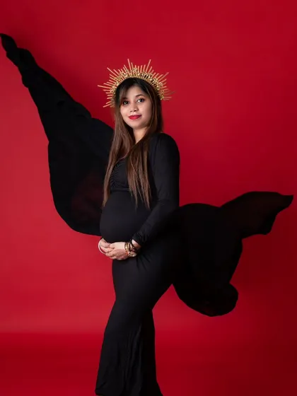A regal maternity portrait of a woman in a black gown and a golden sunburst crown. The bold red background contrasts with the black dress, creating a powerful and queenly image.