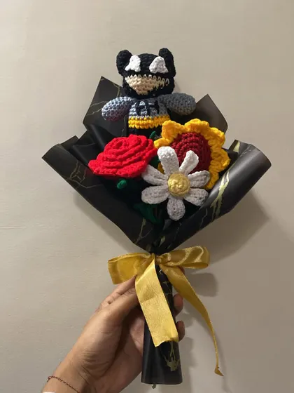 A unique bouquet featuring a Batman-inspired character. The dark wrapping paper with gold accents makes the colors of the crochet rose, sunflower, and daisy pop.