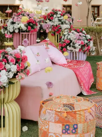 A fun and colorful Mehendi seating area with patchwork ottomans, a pink sofa, and floral arrangements hanging from a tree.