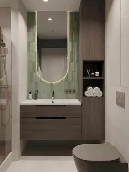 A compact bathroom design featuring a stylish, vertically-tiled green accent wall behind an oval back-lit mirror. The tall storage cabinet next to the vanity provides ample space for all essentials.