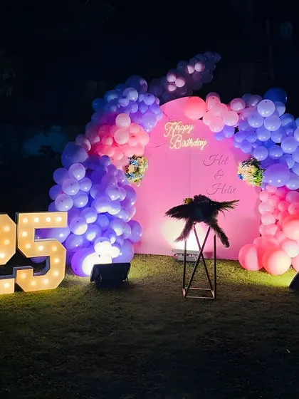 An outdoor 25th birthday celebration at night. The pink backdrop and balloon arch are illuminated by spotlights and the glow of the large marquee numbers.