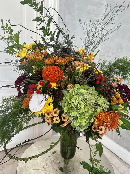 Crisp autumn colours. This arrangement features mustard yellow, pumpkin orange, olive green, and ruby reds, with wild woody stems and foliage for a rustic, seasonal feel.