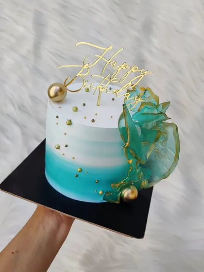 A masterpiece of texture and color. This cake features a teal ombre effect, gold sprinkles, and abstract green rice paper sails.