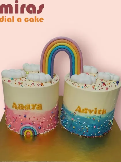 A creative and sweet cake for twins, designed as two separate cakes connected by a rainbow. The pink and blue themes for each twin make it a perfect shared celebration.