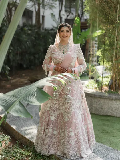A smiling portrait of the bride in her pastel pink lehenga, surrounded by lush greenery.