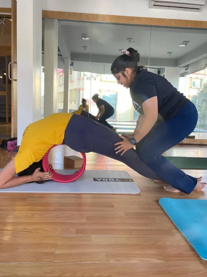 An instructor assists a student in a deep stretch over the yoga wheel, providing support to open the back and shoulders.