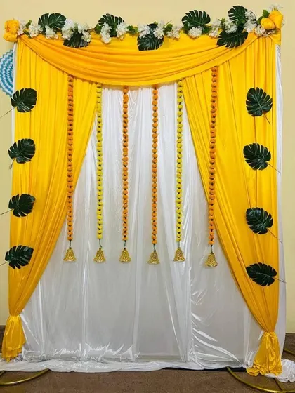 An elegant backdrop with yellow and white drapes, decorated with hanging bells and monstera leaves for a modern tropical feel.