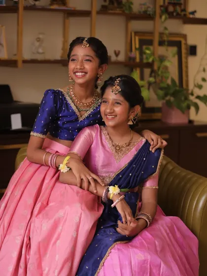Sisters sharing a happy moment in their coordinated traditional outfits. One wears a pink langa dhavani, the other a matching lehenga.