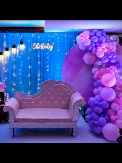 A chic and dreamy baby shower setup. The decor features a purple circular backdrop, a cascading balloon garland in shades of purple, and a fairy light curtain for a magical 'Oh Baby' moment.