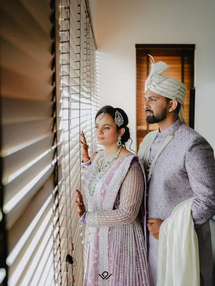 A couple in lavender wedding attire looks out a window, the light creating a dramatic and stylish portrait.