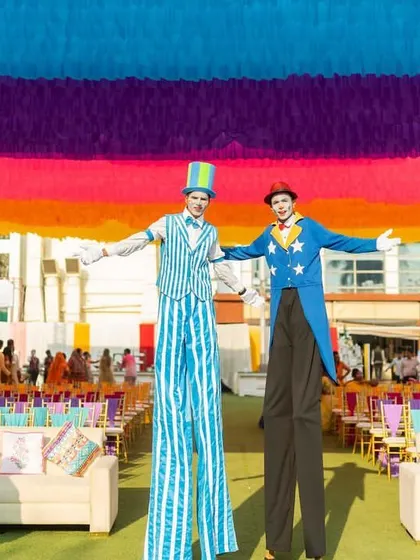 Stilt walkers tower over the colorful seating area, adding to the larger-than-life atmosphere of this vibrant outdoor carnival.