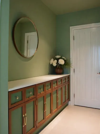 I love creating beautiful vignettes in small corners. This hallway features a custom wooden crockery unit with a white countertop, set against a calming sage green wall with a simple round mirror.