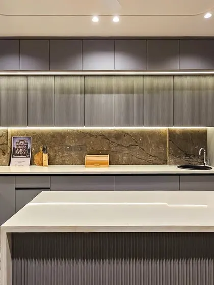 Another angle of the sleek grey kitchen, showing the island and the seamless, handle-less cabinetry. The warm lighting and natural stone backsplash prevent the grey palette from feeling cold.