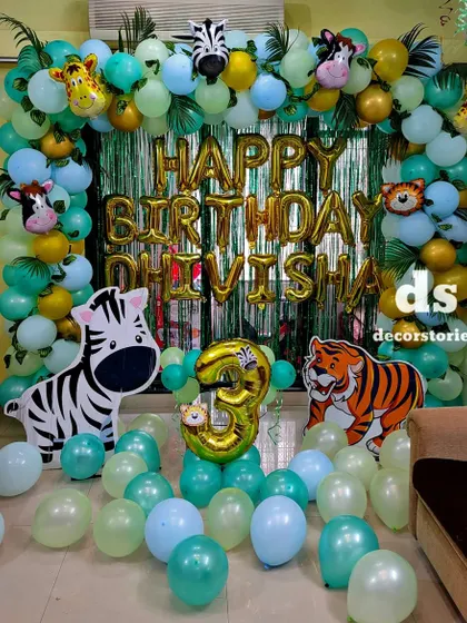 A vibrant jungle theme birthday decoration for a third birthday. This setup features a 'Happy Birthday' foil balloon banner against a shimmering curtain, framed by a lush balloon arch and animal cutouts like a zebra and tiger.