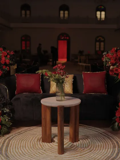 A cozy lounge seating area at the Sangeet, featuring a plush sofa with red and gold cushions, surrounded by dramatic red floral arrangements.