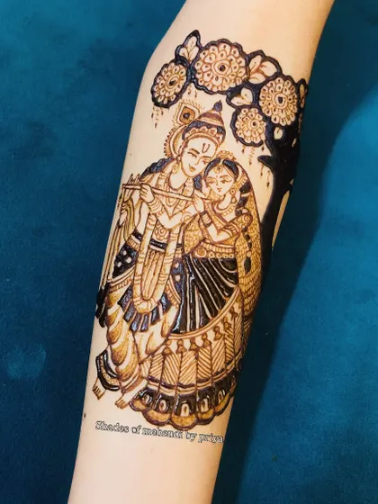 A classic Radha-Krishna figure design on the forearm, standing under a tree. This is a timeless and popular choice for both bridal and festive mehendi.