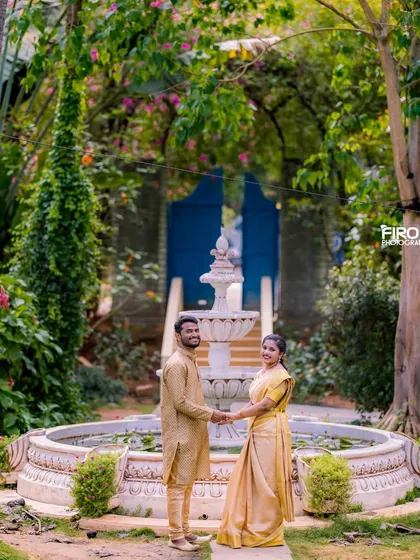 A beautiful full-length portrait of a couple holding hands by a fountain. Their matching golden-yellow outfits look radiant in the lush garden.