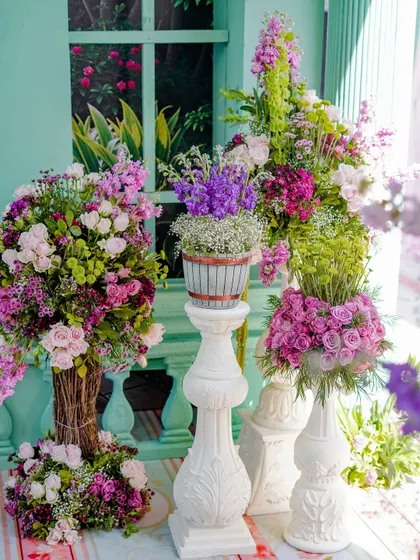 A stunning display of floral arrangements in shades of pink, purple, and white, set against a mint-green wall. The variety of flowers and vases creates a rich and textured visual.