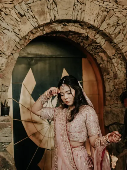 A candid, in-between moment. The bride adjusts her maang tikka, lost in thought, framed by a rustic stone archway that adds a touch of old-world charm.