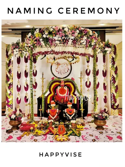 This is a full view of a traditional naming ceremony setup. The decor is rich with floral garlands, brass lamps, and religious idols, creating a truly authentic and blessed environment.