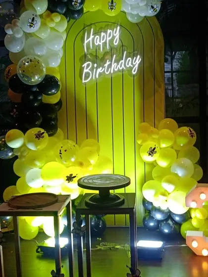 A 'bee-day' party with a honeybee theme, using yellow and black balloons, a honeycomb-style backdrop, and a neon sign.