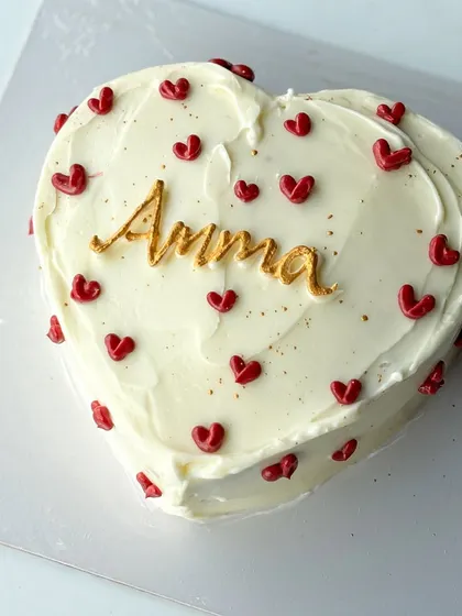 A simple and heartfelt cake for Amma. This heart-shaped cake is covered in white buttercream and decorated with tiny red hearts and a beautiful gold "Amma" script.