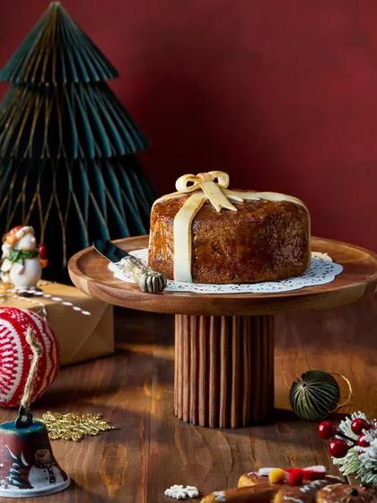 A simple yet elegant shot of a Christmas cake wrapped with a bow, placed on a wooden cake stand. The deep red and green background and festive ornaments create a rich, classic holiday image for Marriott Bonvoy.