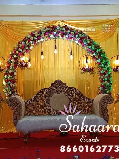 A vibrant reception stage with a floral arch against a bright yellow backdrop. The hanging lanterns and ornate sofa create a festive and traditional setting for any celebration.