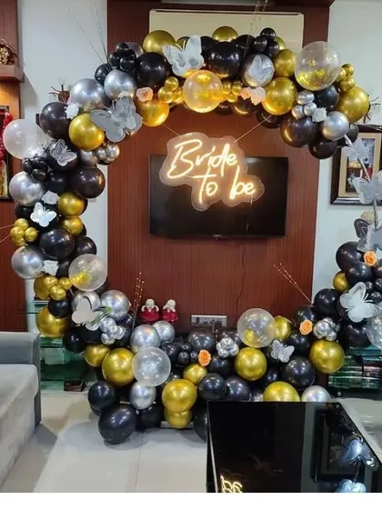 This is a sophisticated black, gold, and silver balloon arch I set up in a living room for a bridal shower. The 'Bride to be' neon sign in the center creates a stylish focal point for the celebration.