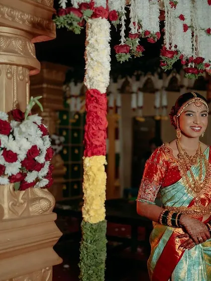 Bhushitha standing by the traditional wedding pillars. Her look is a perfect example of a classic Kannadiga bride, complete with traditional jewelry and a neat bun.
