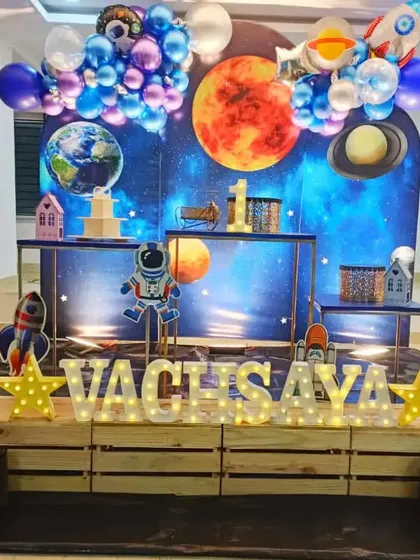 A cosmic first birthday for Vathsaya. This space-themed setup features a backdrop of planets, a cuddly teddy bear astronaut, and light-up letters for a stellar celebration.