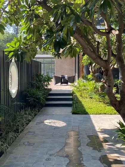 A tranquil pathway at a New Delhi farmhouse, shaded by a mature Plumeria tree. The path leads to a secluded seating area, demonstrating our approach to creating journey and discovery within a garden.