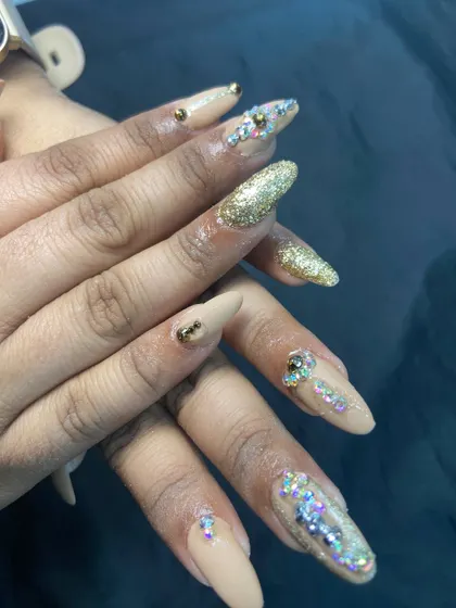 For when you want to make a statement. These long, almond-shaped nail extensions feature a mix of nude polish, dazzling gold glitter, and carefully placed rhinestones for a truly luxurious and festive feel.