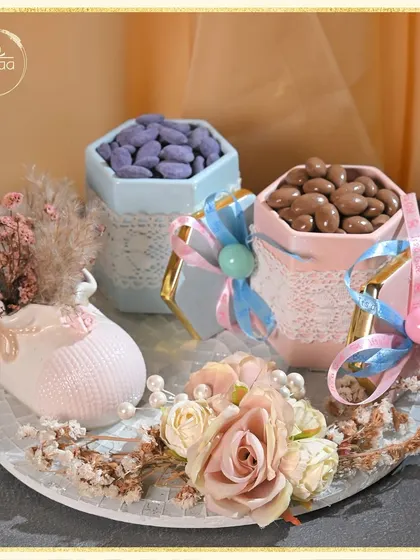 This gender reveal hamper is designed with both pink and blue elements. It features a ceramic baby shoe vase, dried flowers, and jars of pink and blue candies, making it a fun and stylish way to share the surprise.