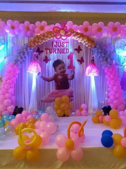 A classic princess-themed first birthday party. The stage is decorated with pink balloon columns and arches, princess cutouts, and a personalized photo of the birthday girl.