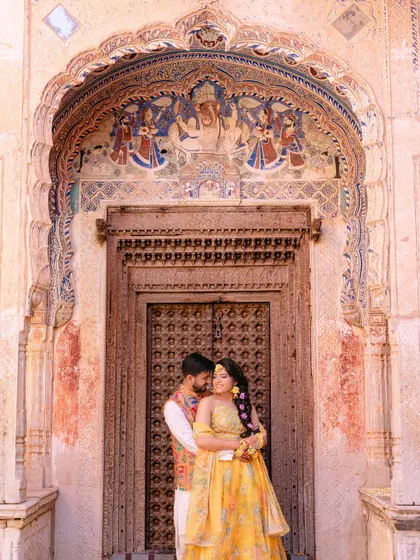The couple embraces before a magnificent, intricately carved wooden door and archway, their vibrant yellow outfits a beautiful contrast to the historic setting.