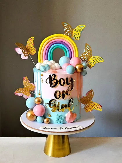A vibrant and festive "Boy or Girl" gender reveal cake. The cake features a pink and blue drip design, a fondant rainbow, and is adorned with golden butterflies and colorful spheres.