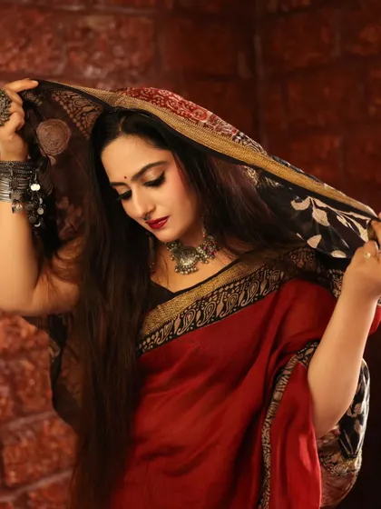 An elegant portrait of a model in a red and black saree. The way she holds the pallu over her head creates a sense of mystery and traditional grace.
