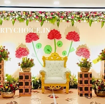 A creative and modern stage for a half saree ceremony. The backdrop features stylized paper flowers and a royal throne, framed by traditional floral pillars.