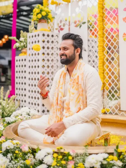A happy groom-to-be, seated amidst a beautiful Haldi setup with a modern jaali backdrop and fresh floral arrangements.