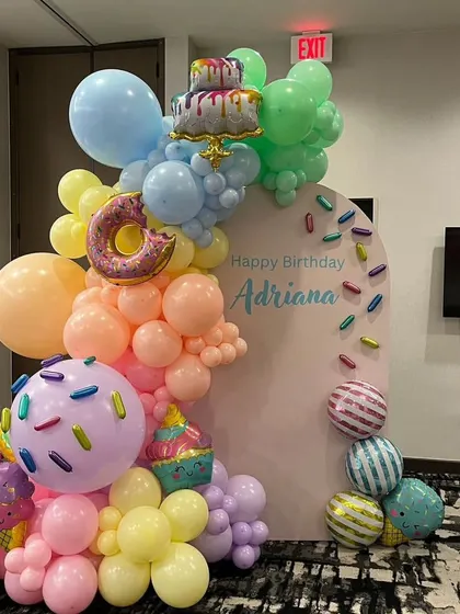 A sweet-treats themed birthday party. This setup features a pastel balloon arch with donut, ice cream, and cake-themed balloons against a custom backdrop.
