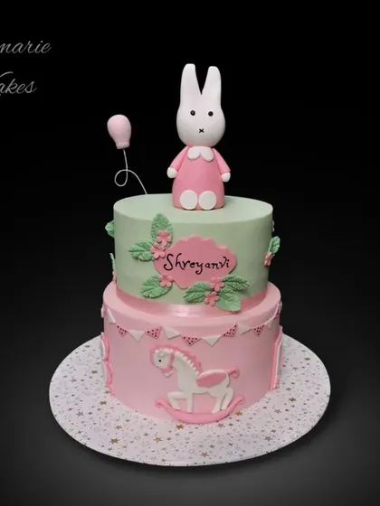 A sweet two-tiered cake for a baby's celebration. It features a pastel pink and green design with a cute bunny topper, a rocking horse detail, and delicate floral accents.