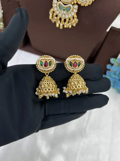 The Tishyaa Gold & Gems necklace. This piece showcases beautiful green Meenakari work in a crescent shape with a detailed pendant and matching jhumkis, all in a rich antique gold finish.