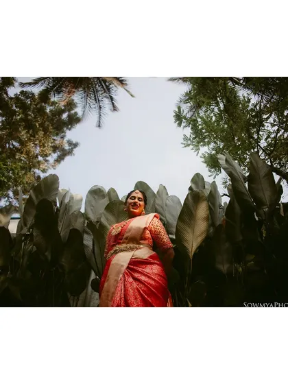 A low-angle shot of a bride in a red saree, surrounded by lush, oversized leaves, creating a dramatic and tropical feel.